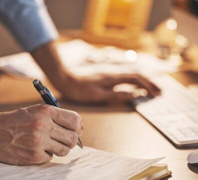 Person writing notes in a notebook while working at a desktop computer