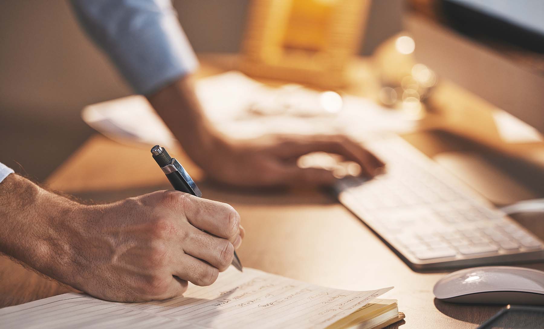 Person writing notes in a notebook while working at a desktop computer